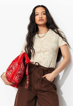 Femme aux longs cheveux foncés portant un top en dentelle crème, un pantalon marron taille haute, un collier chaîne doré, et tenant un sac en tissu rouge à motif cachemire.
