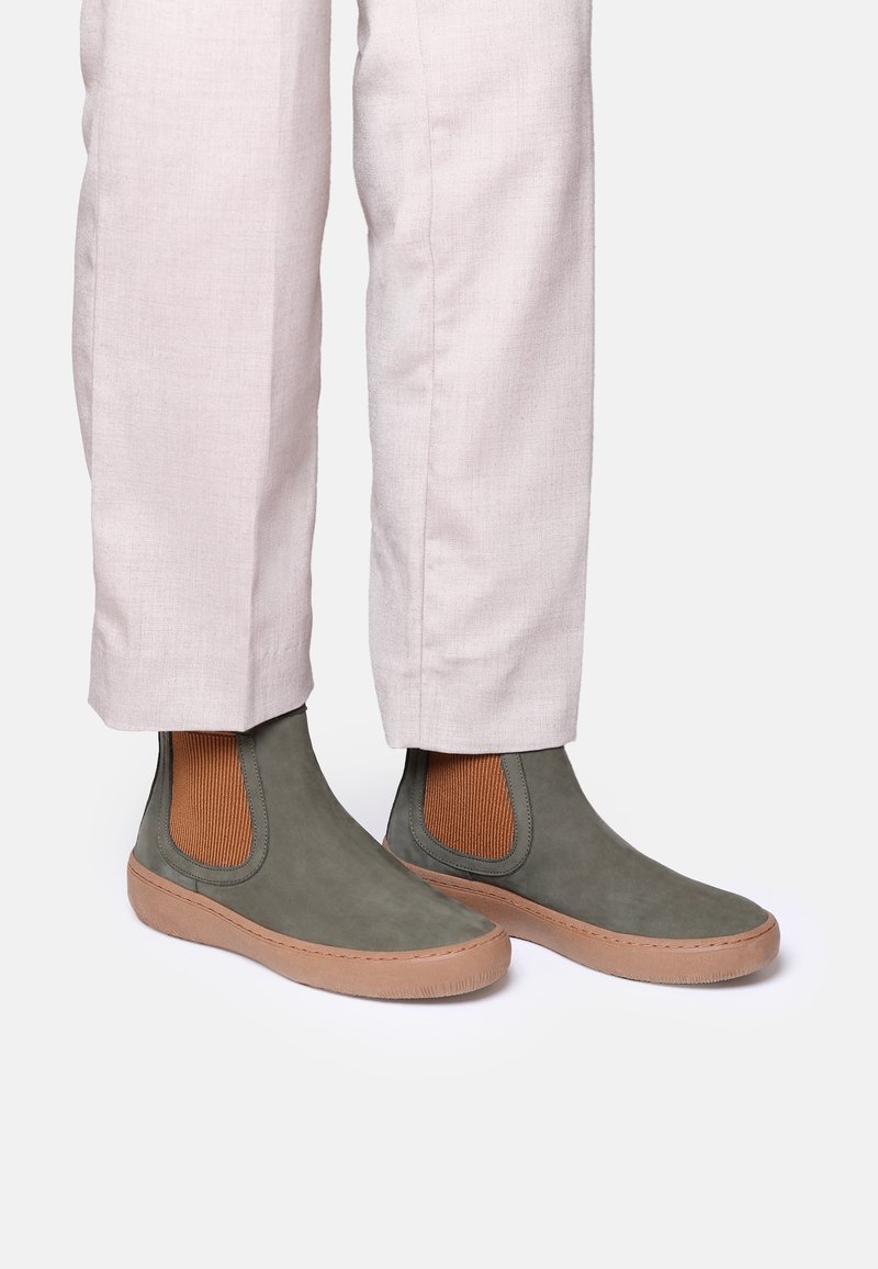 Bottines chelsea en suede vert avec des panneaux élastiques latéraux marron et des semelles en caoutchouc marron clair, assorties à un pantalon ajusté de couleur claire.