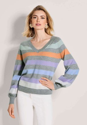 Frau mit blondem Haar, die einen grauen V-Ausschnitt-Pullover mit orangefarbenen, violetten, blauen und weißen horizontalen Streifen trägt, kombiniert mit weißen Hosen und großen Ohrringen.