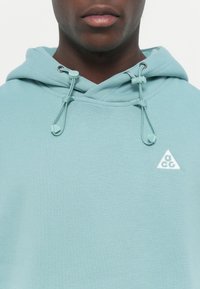 Fleece azul claro feito de um tecido macio, apresentando um logótipo triangular em branco e cordões ajustáveis na capuz. Detalhe de superfície texturizada.