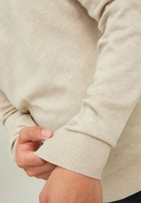 Suéter beige con puños acanalados, textura suave y diseño entallado. Una mano ajusta la manga, destacando su tejido suave.