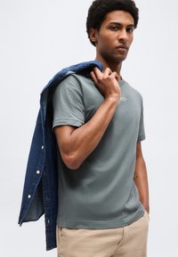 Άνδρας που φορά γκρι μπλουζάκι και μπεζ παντελόνι, κρατώντας ένα μπλε τζιν μπουφάν στον ώμο του, κοιτά λίγο προς τα πλάγια μπροστά σε απλό φόντο.