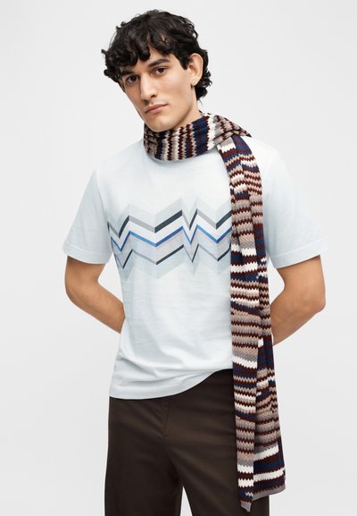 Ανοιχτό μπλε μπλουζάκι με γεωμετρικό σχέδιο σε γκρι, μπλε και μαύρο χρώμα· συνδυασμένο με πολύχρωμο πλεκτό κασκόλ σε κόκκινο, μπεζ και μπλε.