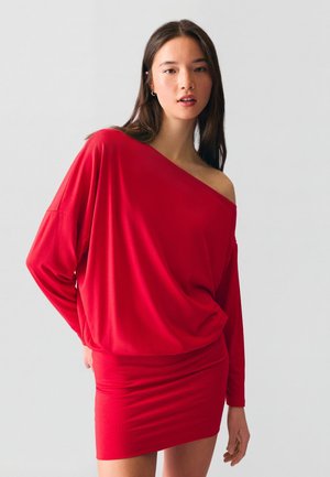 Fiatal nő laza, piros, váll nélküli hosszú ujjú felsőt és hozzáillő, testhezálló miniszoknyát visel, sima világos háttér előtt állva.