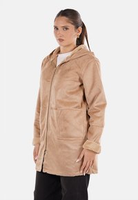 Cappotto in pelliccia sintetica beige con zip frontale, cappuccio, due tasche frontali e polsini foderati in pelliccia. Presenta una texture liscia e una vestibilità comoda.