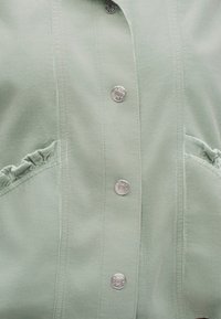 Gros plan sur un tissu vert clair avec des coutures verticales, des boutons-pression métalliques et un petit luffle froncé près des bords des poches.