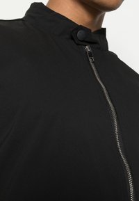 Veste noire avec un col haut, une fermeture éclair à l'avant et un bouton-pression au col. Le matériau semble lisse avec une finition mate.