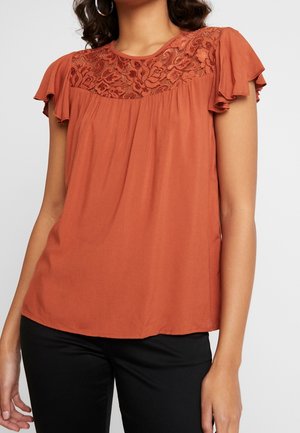 Vrouw met een roestoranje blouse met kantdetail op de bovenborst en vlinderachtige mouwen, gecombineerd met een zwarte broek.