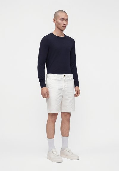 Homme debout, portant une chemise noire à manches longues, un short blanc longueur genoux, des chaussettes blanches et des baskets blanches basses sur fond uni.