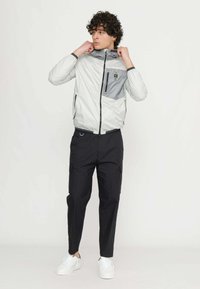 Let vægt, grå lynlåsjakke med en grå lomme, sorte accenter og hætte; båret med sorte cargo-bukser og hvide sneakers.