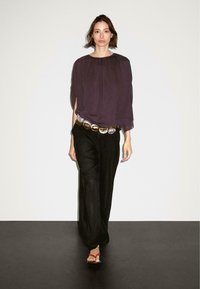 Top viola largo, pantaloni neri a gamba larga, cintura con dettagli dorati e sandali rossi, su una modella contro uno sfondo chiaro.
