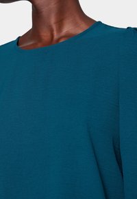 Indumento teal con un tessuto liscio e testurizzato. Presenta un collo rotondo e morbide pieghe sulle spalle, evidenziando una vestibilità rilassata.