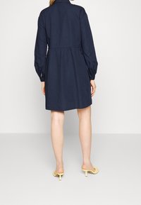 Marinblå skjortklänning med långa puffärmar, midjeanpassad och raka fållar. Tyget verkar lätt och jämnt. Bärs med gula klacksandaler.