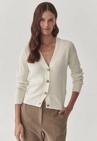 Cremfarbener gestrickter Cardigan mit tiefem V-Ausschnitt, geripptem Textur, fünf Holzknöpfen und langen Ärmeln. Kombiniert mit braunen Hosen.