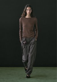 Jeune femme aux longs cheveux ondulés marchant, portant un haut à manches longues rayé marron, un pantalon ample foncé, une petite pochette suspendue, et des chaussures noires.