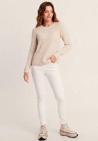 Pull en tricot beige avec des motifs texturés, des manches longues et des boutons aux poignets, associé à un pantalon blanc ajusté et des chaussures beiges.