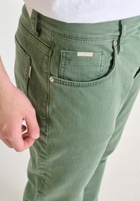 Pantalones de algodón verdes con un acabado texturizado, que incluyen un bolsillo delantero, una etiqueta de marca y un detalle de botón en el costado.