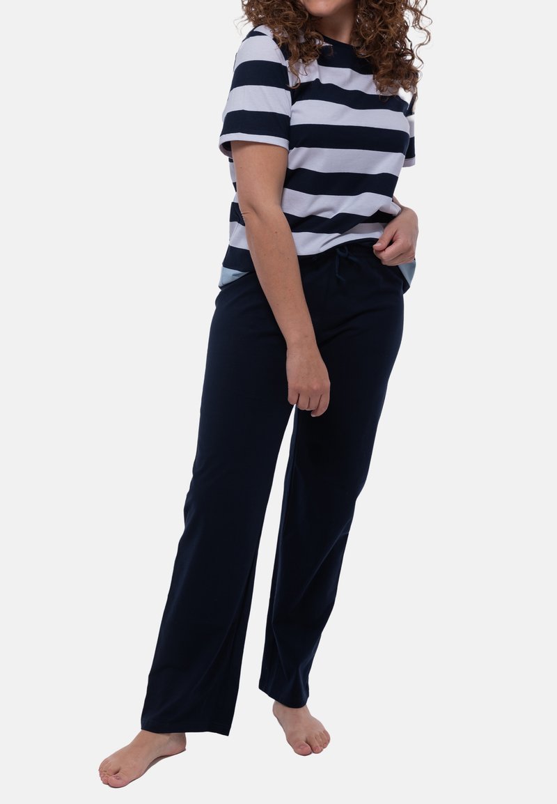 Kobieta z kręconymi włosami, ubrana w koszulę w czarno-białe paski z krótkim rękawem oraz luźne ciemne spodnie, stojąca boso na białym tle.