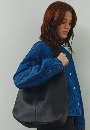 Sac hobo en cuir noir avec une surface texturée. Forme douce et arrondie. Porté sur l'épaule, associé à une veste bleue matelassée.