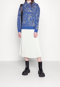 Felpa blu con motivo paisley, gonna plissettata bianca e stivali chunky neri con tacco. La felpa presenta un cappuccio con cordoncino e polsini a coste.