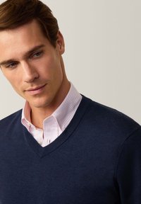 Maglione blu navy a scollo a V sovrapposto a una camicia a righe light pink e bianca. Tessuto liscio, design aderente e collettino classico.