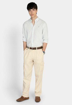 Jeune homme debout les mains dans les poches, portant une chemise légère rayée à boutons, un pantalon cargo beige, une ceinture marron et des mocassins marron.