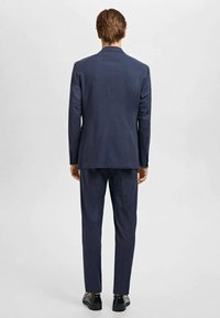Costume marine foncé avec un design ajusté, présentant une fente simple à l'arrière, un pantalon slim et des chaussures noires brillantes.
