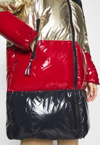 Puffjacka med guld-, röd- och marinblå sektioner med glansiga ytor. Inkluderar en ficka med dragkedja och ett horisontellt färgblockeringsdesign.