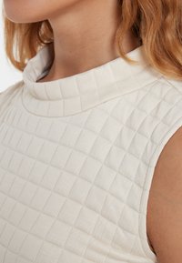 Cremefarbenes Oberteil mit einem strukturierten, gesteppten Muster, einem hohen, abgerundeten Kragen und ärmellosem Design, hergestellt aus weichem, dehnbarem Stoff.
