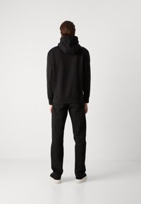 Modelo masculino de pie, de espaldas, vistiendo una sudadera con capucha negra, jeans negros y zapatillas en blanco y negro contra un fondo blanco sencillo.