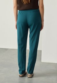 Pantalones color verde azulado hechos de una tela suave, con un diseño de pierna recta y un solo bolsillo trasero, combinados con zapatos estampados.