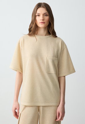 Chemise beige à manches courtes en tissu doux et texturé. Elle présente une poche sur la poitrine gauche et une coupe décontractée.