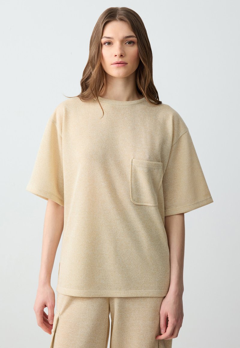 Chemise beige à manches courtes en tissu doux et texturé. Elle présente une poche sur la poitrine gauche et une coupe décontractée.