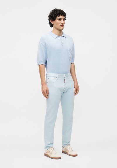 Νέος άνδρας στέκεται, φοράει ανοιχτό μπλε πόλο με κοντά μανίκια, ανοιχτά μπλε τζιν και μπεζ αθλητικά παπούτσια σε λευκό φόντο.
