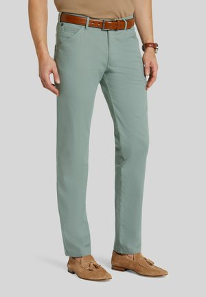 Mann trägt hellgrüne Slim-Fit-Hose, braunen Ledergürtel, beige Tassel-Loafer und eine braune Uhr vor grauem Hintergrund.