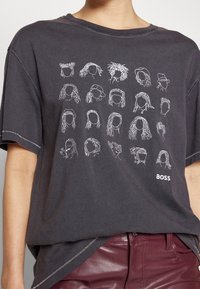 Mörkgrå T-shirt med vit linjekonst av 20 olika ansiktslösa frisyrer, "BOSS"-texten på nedre högra sidan, bärs tillsammans med mörkröda läderbyxor.
