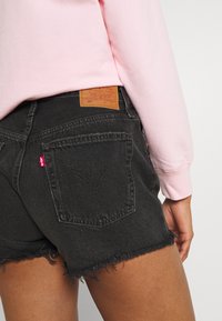 Svarta denimshorts med fransade kanter, med en brun läderlapp på ryggen. Logotyp med röd etikett synlig på fickan.