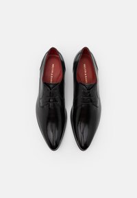 Scarpe da dress in pelle nera con punta affusolata, finitura lucida, design con lacci e fodera interna rossa con testo dorato.