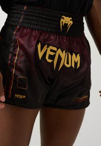 Crne in burgundske kratke hlače iz lahkega materiala, ki imajo v zlati barvi napis "Venum", rdečimi detajli in elastičnim pasom.