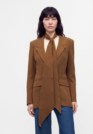 Frau mit langen schwarzen Haaren trägt einen braunen Blazer mit großem Schal und blaue Jeans, steht vor einem schlichten hellen Hintergrund.