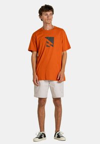 Camiseta naranja de manga corta con un diseño gráfico negro en el pecho, combinada con pantalones cortos beige claros y zapatillas negras y blancas.