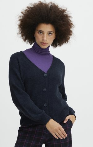 Cardigan bleu marine foncé en tricot texturé, avec un col en V, des manches longues et quatre grands boutons. Porté par-dessus un col roulé violet.