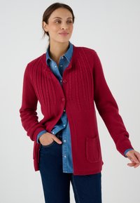 Cárdigan de punto burdeos con patrones verticales texturizados, cierre de botones y un bolsillo. Superpuesto sobre una camisa de denim y jeans oscuros.