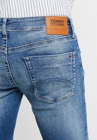 Zadní pohled na osobu, která má na sobě světle modré džíny Tommy Jeans s koženou nášivkou značky a malým vlajkovým logem na zadní kapse.