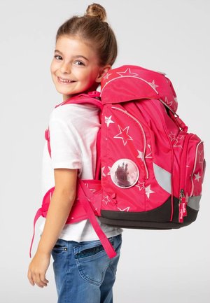 Ein lächelndes junges Mädchen trägt ein weißes Shirt und eine blaue Jeans und hält einen großen rosa Rucksack mit Sternen-Muster und einem Schloss-Abzeichen.