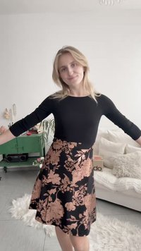 Žena v černém topu a květované sukni stojí s rozpaženýma rukama v obývacím pokoji se zelenou skříňkou a bílou pohovkou.