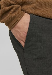 Manschet della maglia lavorata a maglia marrone con finitura testurizzata, vicino a pantaloni grigi scuri con una tasca laterale visibile e un sottile motivo a spina di pesce.