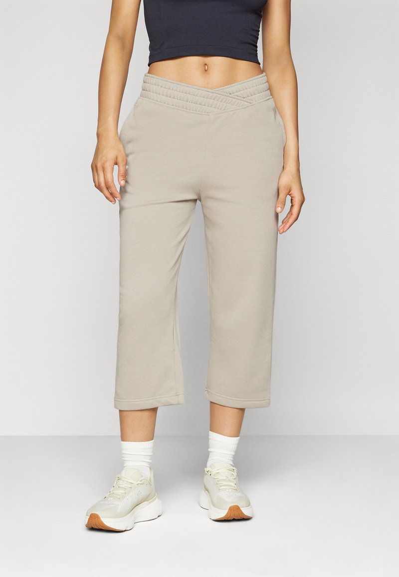 Beige, kurz geschnittene Jogginghose mit einem strukturierten, hochtaillierten Design, ausgestattet mit einem glatten Stoff und umgeschlagenen Säumen, kombiniert mit weißen Sportschuhen.