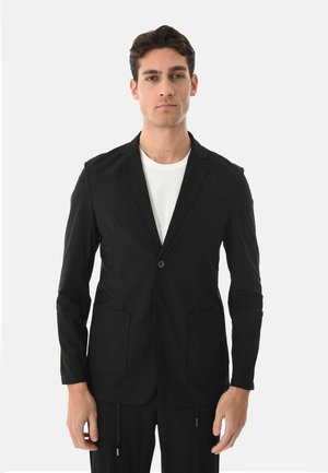 Giovane uomo con blazer nero a un bottone indossato sopra una t-shirt bianca, in piedi davanti a uno sfondo grigio chiaro uniforme.