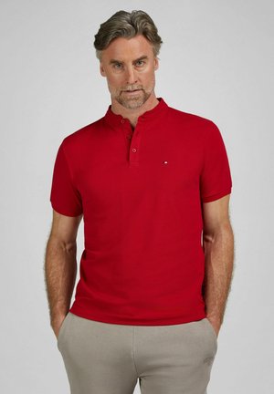 Mand med gråt hår og skæg iført rød kortærmet henley-shirt og beige bukser, stående med hænderne i lommerne mod en ensfarvet baggrund.